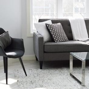 Modern living room with gray sofa, black chair, glass coffee table, and patterned cushions on a light rug near large windows.
