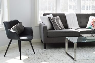 Modern living room with gray sofa, black chair, glass coffee table, and patterned cushions on a light rug near large windows.