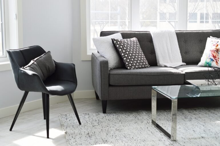 Modern living room with gray sofa, black chair, glass coffee table, and patterned cushions on a light rug near large windows.