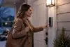 A woman in a brown coat rings a doorbell camera at dusk, holding a large handbag. The home entrance is warmly lit, creating a welcoming atmosphere.