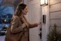 A woman in a brown coat rings a doorbell camera at dusk, holding a large handbag. The home entrance is warmly lit, creating a welcoming atmosphere.