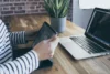Person in striped shirt uses tablet next to an open laptop displaying code on a wooden desk with a potted plant.