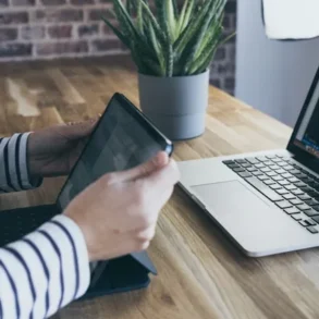 Person in striped shirt uses tablet next to an open laptop displaying code on a wooden desk with a potted plant.