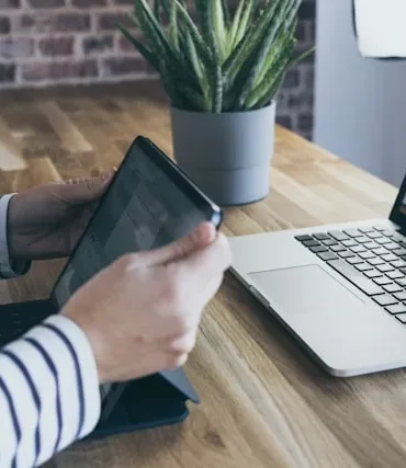 Person in striped shirt uses tablet next to an open laptop displaying code on a wooden desk with a potted plant.