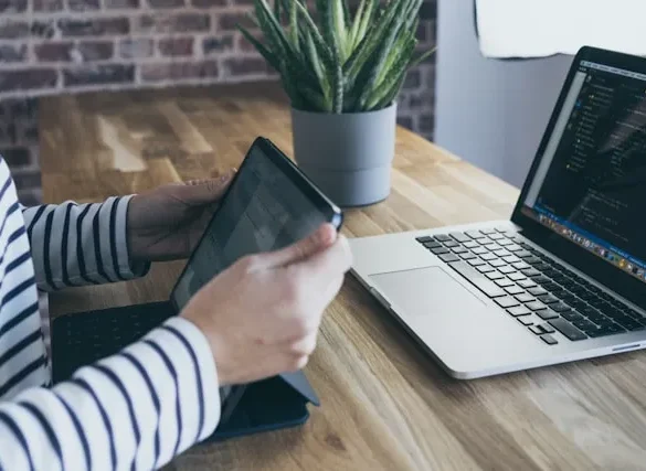 Person in striped shirt uses tablet next to an open laptop displaying code on a wooden desk with a potted plant.