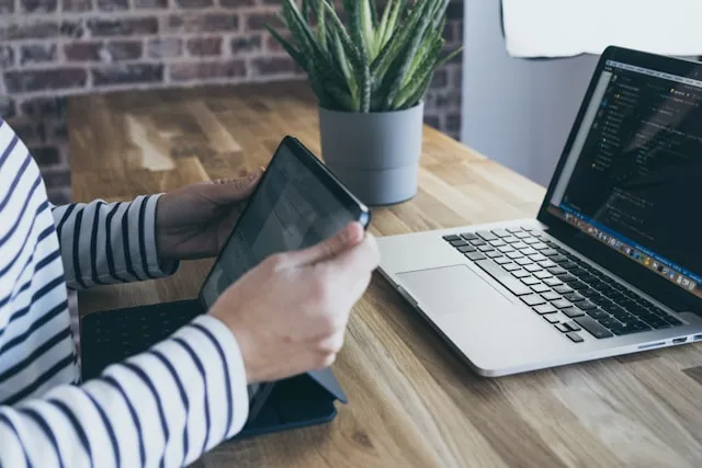 Person in striped shirt uses tablet next to an open laptop displaying code on a wooden desk with a potted plant.