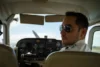 A pilot wearing sunglasses and a uniform seated in the cockpit of a small aircraft, looking back. The cockpit displays various controls and gauges.