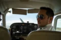 A pilot wearing sunglasses and a uniform seated in the cockpit of a small aircraft, looking back. The cockpit displays various controls and gauges.