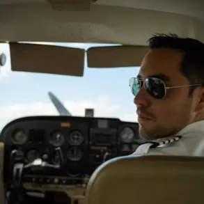 A pilot wearing sunglasses and a uniform seated in the cockpit of a small aircraft, looking back. The cockpit displays various controls and gauges.