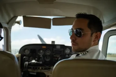 A pilot wearing sunglasses and a uniform seated in the cockpit of a small aircraft, looking back. The cockpit displays various controls and gauges.