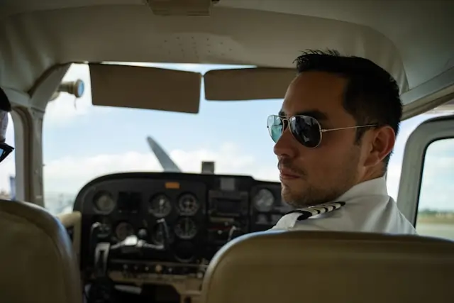A pilot wearing sunglasses and a uniform seated in the cockpit of a small aircraft, looking back. The cockpit displays various controls and gauges.
