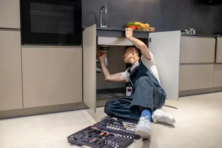 Person in overalls fixing plumbing under a kitchen sink with toolset on the floor nearby.