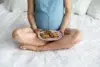 Pregnant woman sitting cross-legged on a bed holding a plate of chocolate chip cookies in her lap.