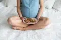 Pregnant woman sitting cross-legged on a bed holding a plate of chocolate chip cookies in her lap.