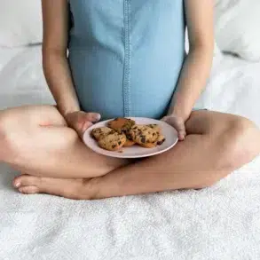 Pregnant woman sitting cross-legged on a bed holding a plate of chocolate chip cookies in her lap.