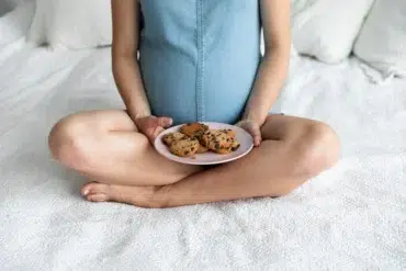 Pregnant woman sitting cross-legged on a bed holding a plate of chocolate chip cookies in her lap.