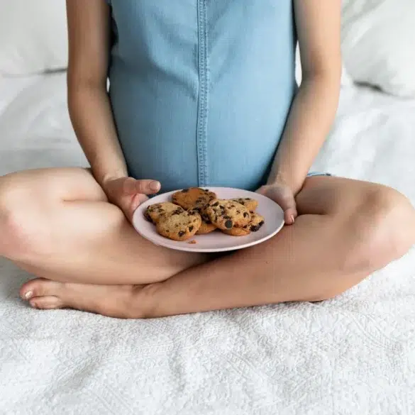 Pregnant woman sitting cross-legged on a bed holding a plate of chocolate chip cookies in her lap.