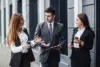 Three business professionals in suits converse outside, each holding coffee cups. The atmosphere is engaged and collaborative, suggesting a lively discussion.
