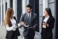 Three business professionals in suits converse outside, each holding coffee cups. The atmosphere is engaged and collaborative, suggesting a lively discussion.