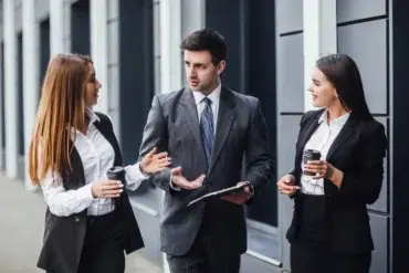 Three business professionals in suits converse outside, each holding coffee cups. The atmosphere is engaged and collaborative, suggesting a lively discussion.