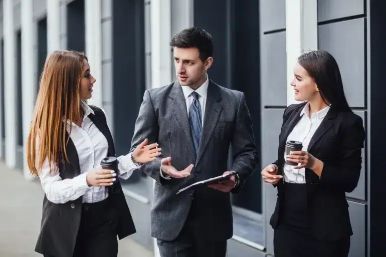 Three business professionals in suits converse outside, each holding coffee cups. The atmosphere is engaged and collaborative, suggesting a lively discussion.