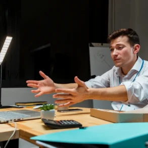 A frustrated businessman gestures while looking at a laptop on his desk in a dimly lit office.
