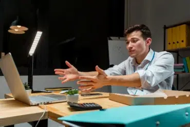 A frustrated businessman gestures while looking at a laptop on his desk in a dimly lit office.