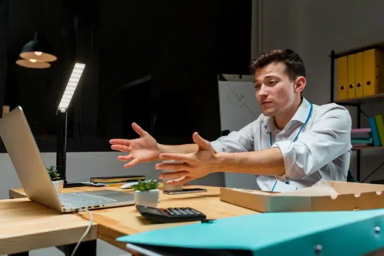 A frustrated businessman gestures while looking at a laptop on his desk in a dimly lit office.