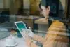 Woman in glasses and a yellow sweater using a tablet next to a cup of coffee at a cafe.