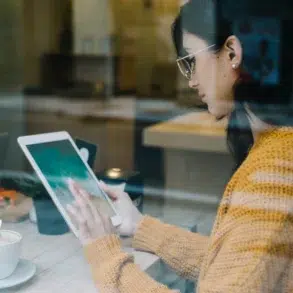 Woman in glasses and a yellow sweater using a tablet next to a cup of coffee at a cafe.