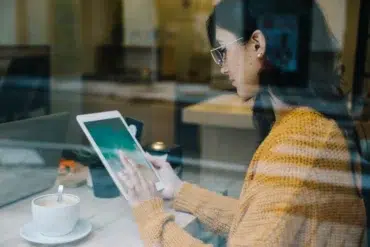 Woman in glasses and a yellow sweater using a tablet next to a cup of coffee at a cafe.