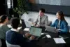 Four people in a modern office setting are gathered around a dark table, engaged in a meeting with laptops and documents.