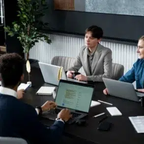 Four people in a modern office setting are gathered around a dark table, engaged in a meeting with laptops and documents.
