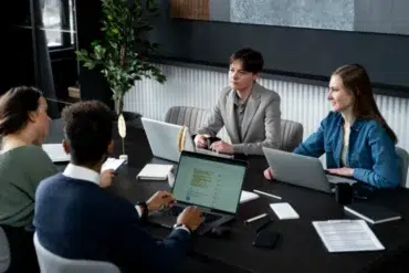 Four people in a modern office setting are gathered around a dark table, engaged in a meeting with laptops and documents.