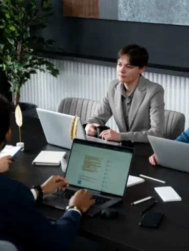 Four people in a modern office setting are gathered around a dark table, engaged in a meeting with laptops and documents.