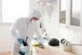 A person in full protective gear including a white hazmat suit, hood, face mask, and goggles wipes down a kitchen counter with a yellow cloth while holding a spray bottle filled with blue liquid.