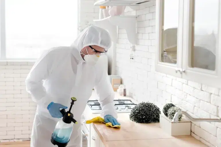 A person in full protective gear including a white hazmat suit, hood, face mask, and goggles wipes down a kitchen counter with a yellow cloth while holding a spray bottle filled with blue liquid.