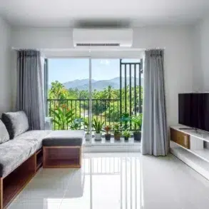 Modern living room with a gray sectional sofa, TV stand, and large sliding glass doors opening to a balcony with lush green mountain views.
