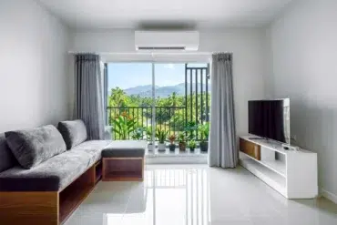 Modern living room with a gray sectional sofa, TV stand, and large sliding glass doors opening to a balcony with lush green mountain views.