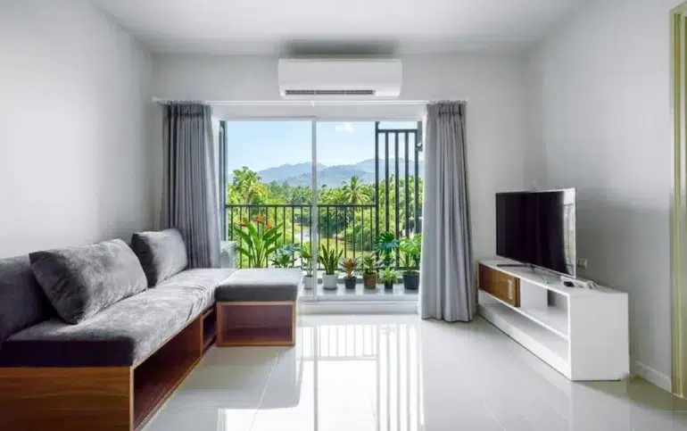Modern living room with a gray sectional sofa, TV stand, and large sliding glass doors opening to a balcony with lush green mountain views.