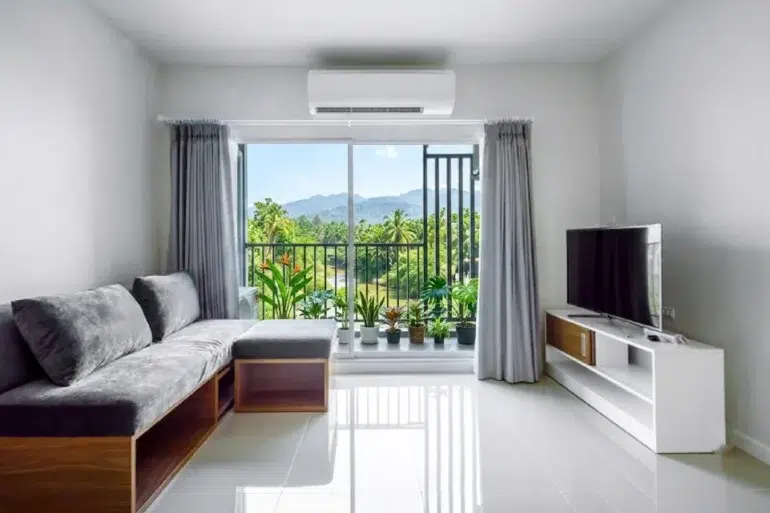 Modern living room with a gray sectional sofa, TV stand, and large sliding glass doors opening to a balcony with lush green mountain views.