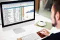 Person in suit works on a computer spreadsheet with charts, a coffee cup nearby.
