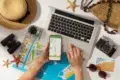 A person planning a trip with a laptop, smartphone with a map, paper maps, camera, hat, and sunglasses on a table. Mood is adventurous and organized.