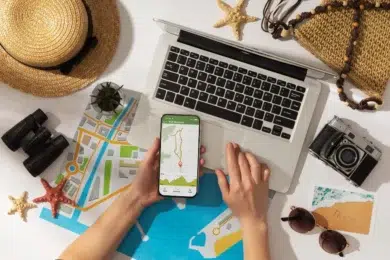 A person planning a trip with a laptop, smartphone with a map, paper maps, camera, hat, and sunglasses on a table. Mood is adventurous and organized.