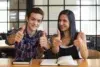 Two smiling young adults give thumbs up while sitting at a table with books and a phone.