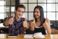 Two smiling young adults give thumbs up while sitting at a table with books and a phone.