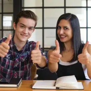 Two smiling young adults give thumbs up while sitting at a table with books and a phone.