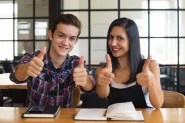 Two smiling young adults give thumbs up while sitting at a table with books and a phone.
