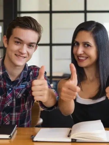 Two smiling young adults give thumbs up while sitting at a table with books and a phone.