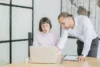 A man in a white shirt leans over a woman seated at a desk to look at her laptop screen in a bright, modern office.